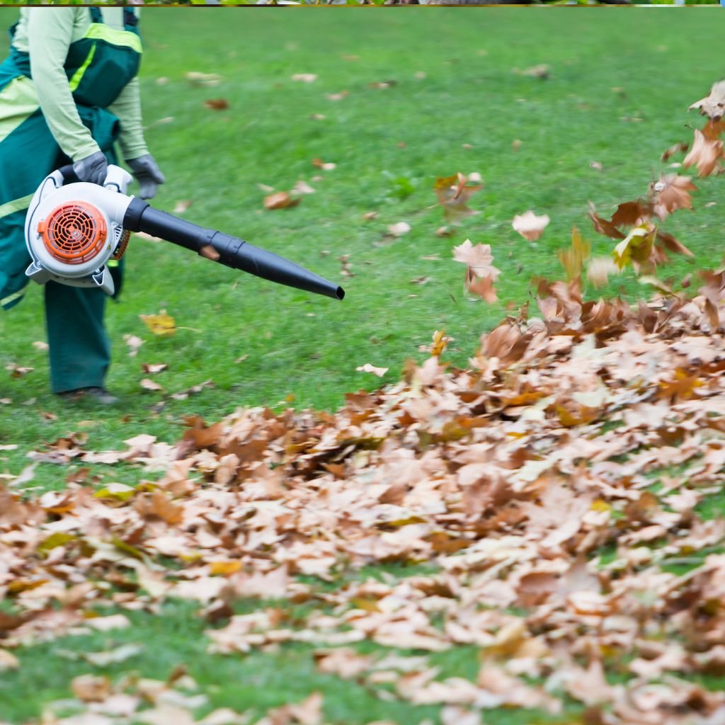 Landscape maintenance professional using a backpack leaf blower to efficiently clear and consolidate a large pile of dry autumn leaves on a green lawn, demonstrating comprehensive yard cleanups and leaf removal services.