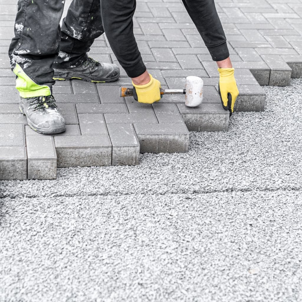 Professional hardscape installer placing interlocking grey pavers in a herringbone pattern on a crushed gravel base, wearing yellow gloves and safety boots, during a pave installation project in Las Vegas.