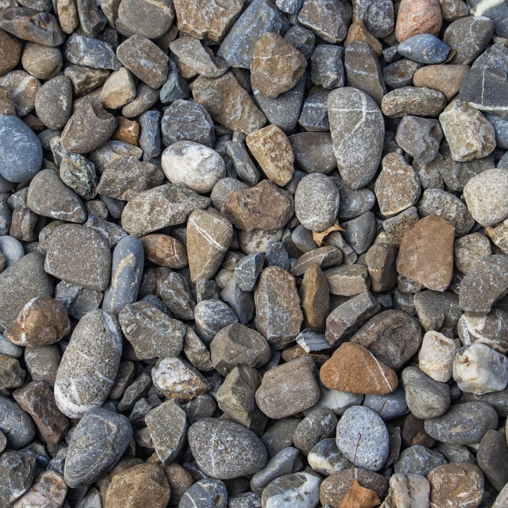 Close-up texture of mixed, multi-colored river gravel and smooth, rounded stones used as a base layer or decorative ground cover for a stone paver installation project.