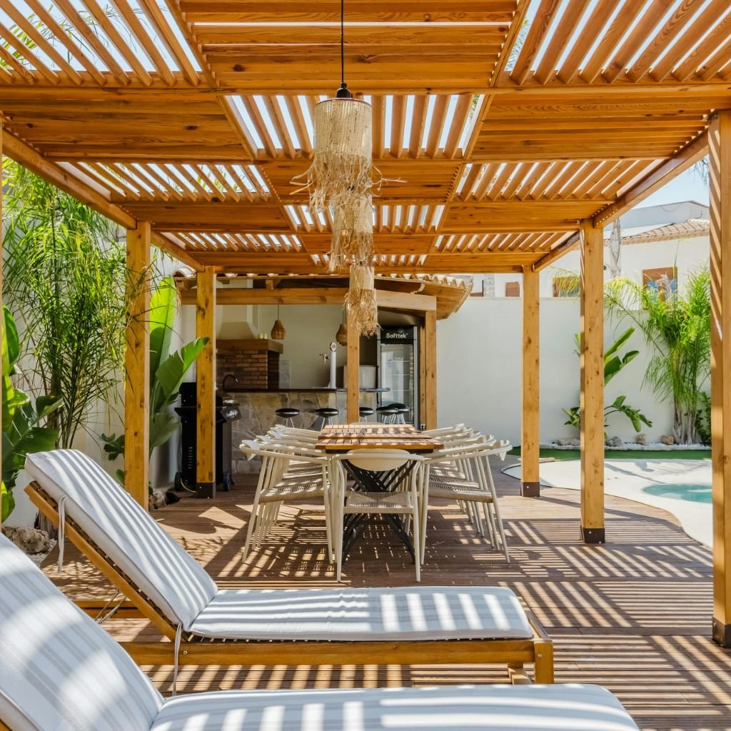 Luxury backyard patio area featuring a custom wooden slatted pergola and patio cover providing shade over a modern outdoor dining set, surrounded by lounge chairs and a pool area, ideal for patio cover installation in a residential setting.